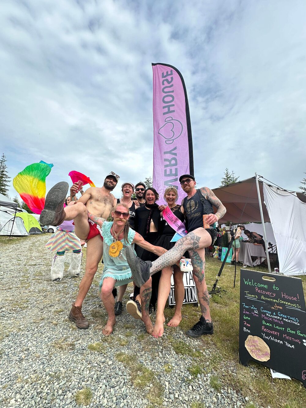 The Recovery House crew posing with legs in the air beside the camp's pink Recovery House flag and a chalkboard welcome sign at Otherworld
