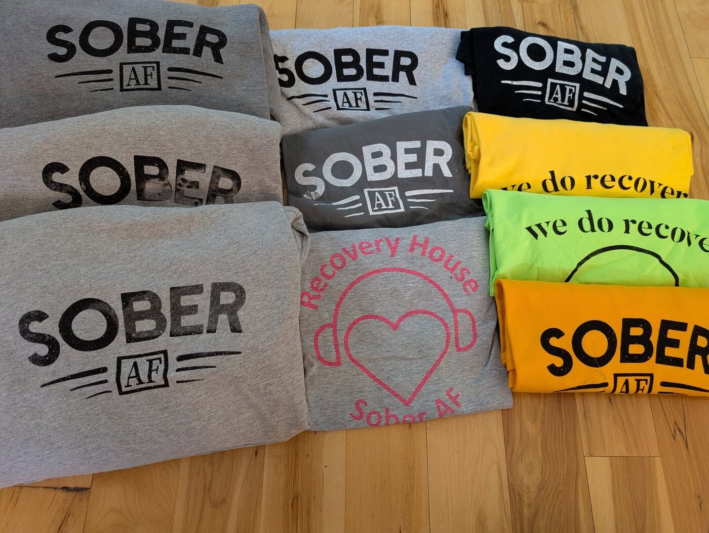 A spread of Recovery House t-shirts in grey, green, yellow and white
