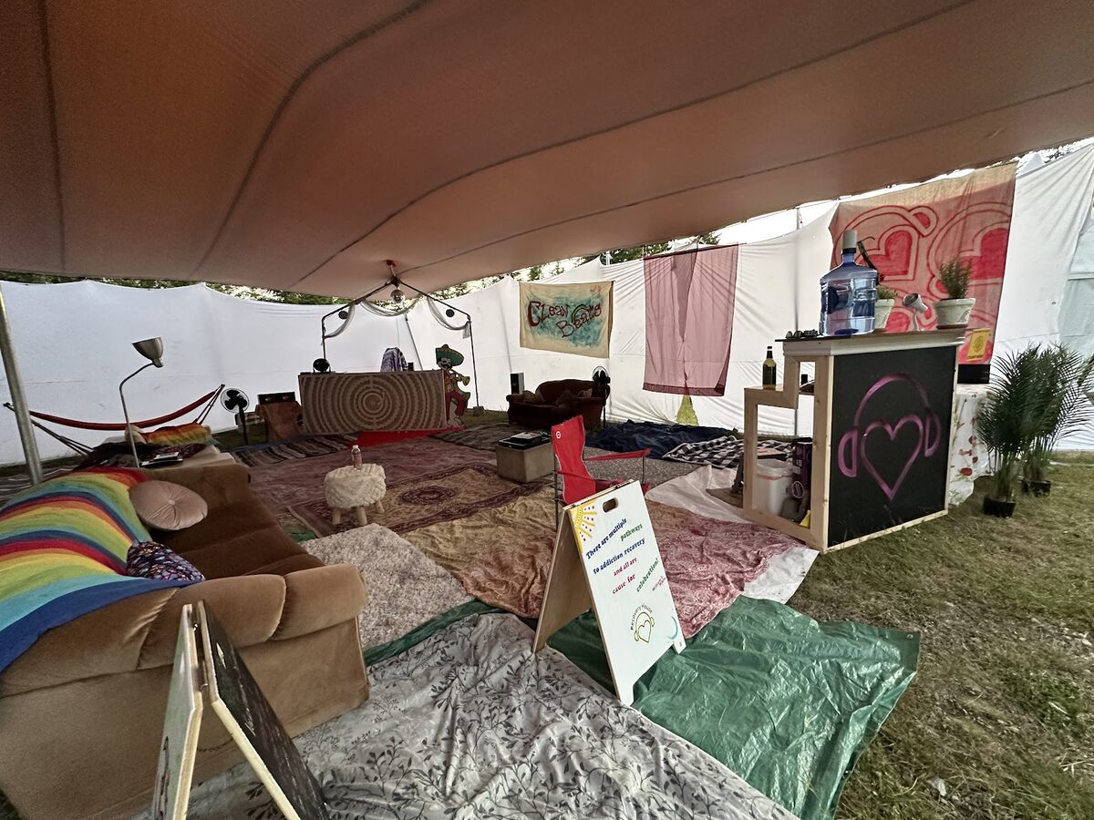 The Recovery House camp with a rainbow couch, sandwich-board signs and a bar painted with the Recovery House logo